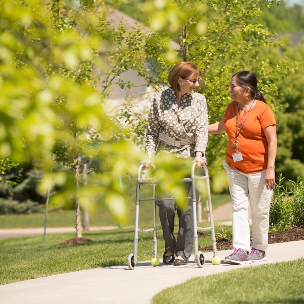 A caregiver warmly supports a smiling resident using a walker on a sunny garden path.