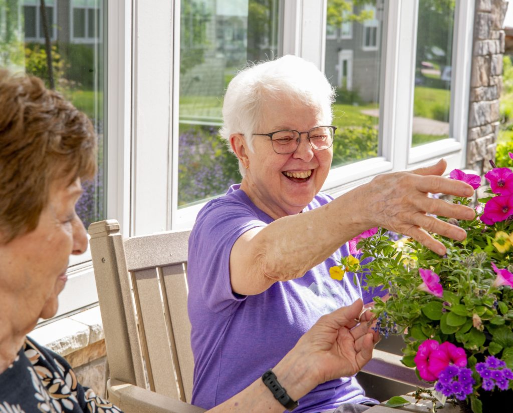Two smiling elderly women enjoying chatting by flowering plants on a bright patio.