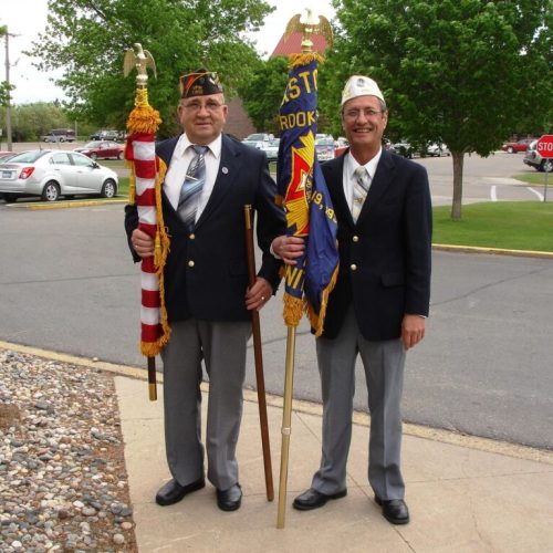 Two smiling veterans proudly holding flags outside on a sunny day, surrounded by greenery.