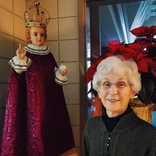 Smiling elderly woman stands beside a decorated statue with vibrant red poinsettias indoors.