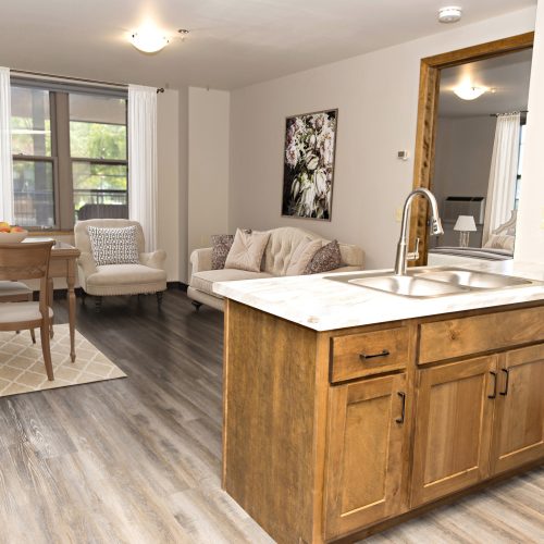Sunlit living and dining area with comfortable seating and warm wooden kitchen cabinetry.