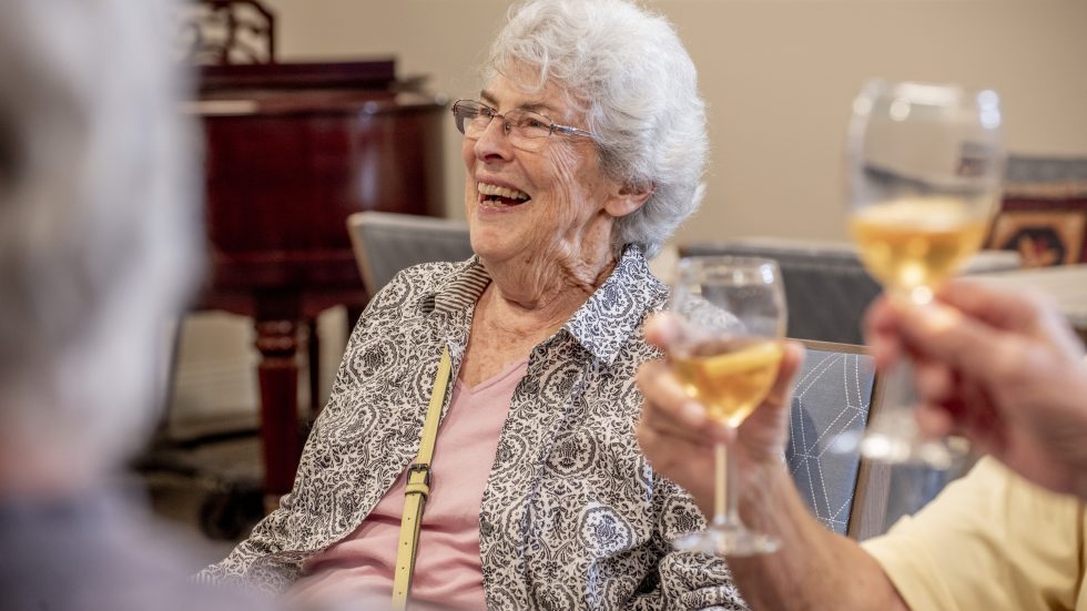 An elderly woman joyfully socializing and raising a glass in a warm, inviting setting.