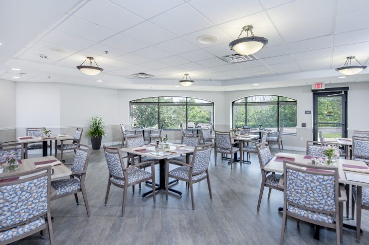 Bright dining room with patterned chairs, tables set for meals, and large windows overlooking greenery.