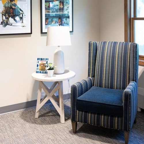 Upholstered armchair beside a table with a lamp and plant, bright room with natural light.