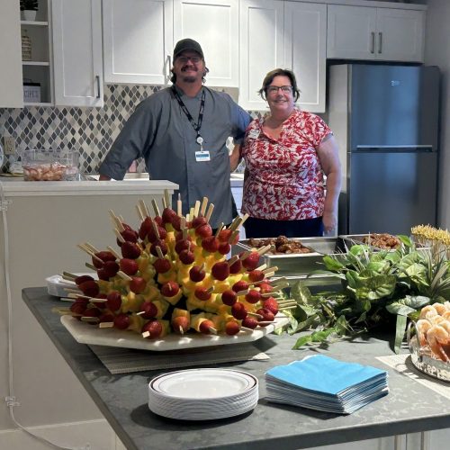 Two smiling staff stand behind a kitchen counter with fresh appetizers and snacks displayed.