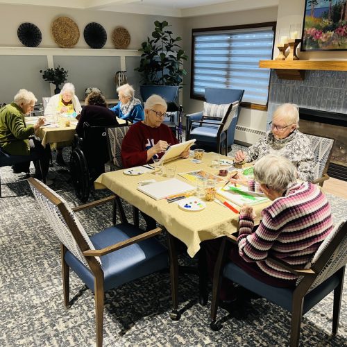 Seniors engaged in a friendly painting activity in a warm, inviting community room.