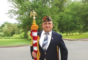 Veteran in uniform holding a ceremonial flag pole in a peaceful, green outdoor setting.