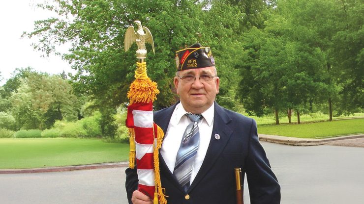 Veteran in uniform holding a ceremonial flag pole in a peaceful, green outdoor setting.