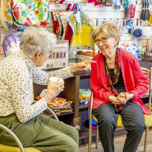 Two elderly women share a joyful conversation in a bright community room filled with colorful crafts and supplies.