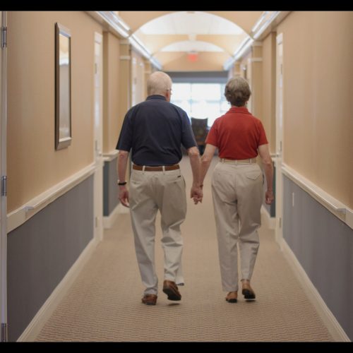 Elderly couple walking hand in hand down a bright, welcoming hallway in a retirement community.