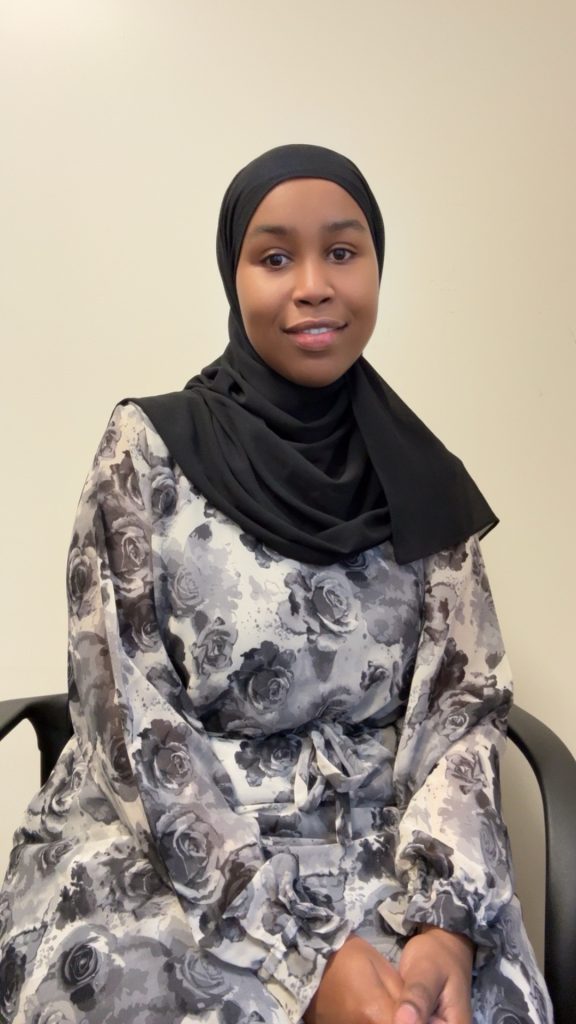 Friendly staff member wearing a floral dress and black hijab, embodying warmth and care.