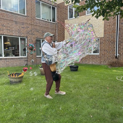 An elderly man joyfully creates giant bubbles in a sunny courtyard surrounded by brick buildings.