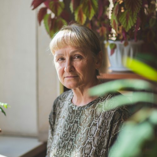 Senior woman sitting near a window surrounded by lush indoor plants in a peaceful space.