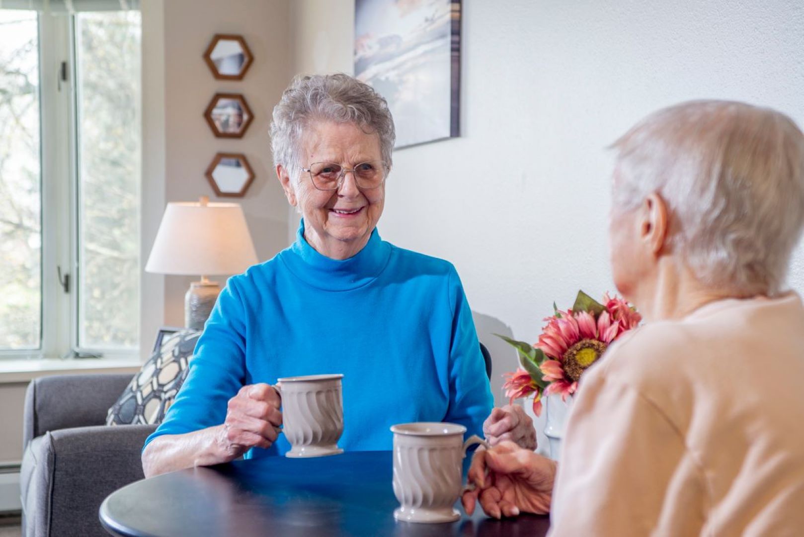 Two senior females sitting together at a table talking and smiling while having coffee.